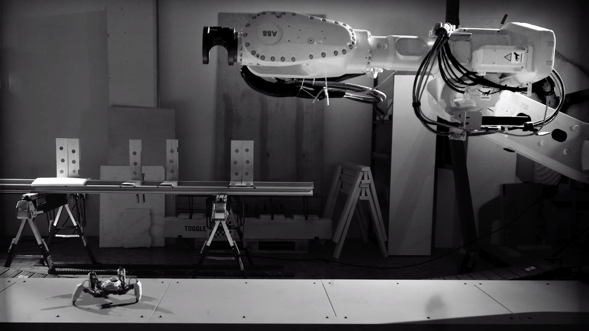 A grayscale video still showing a very small robot approaching a very large robot