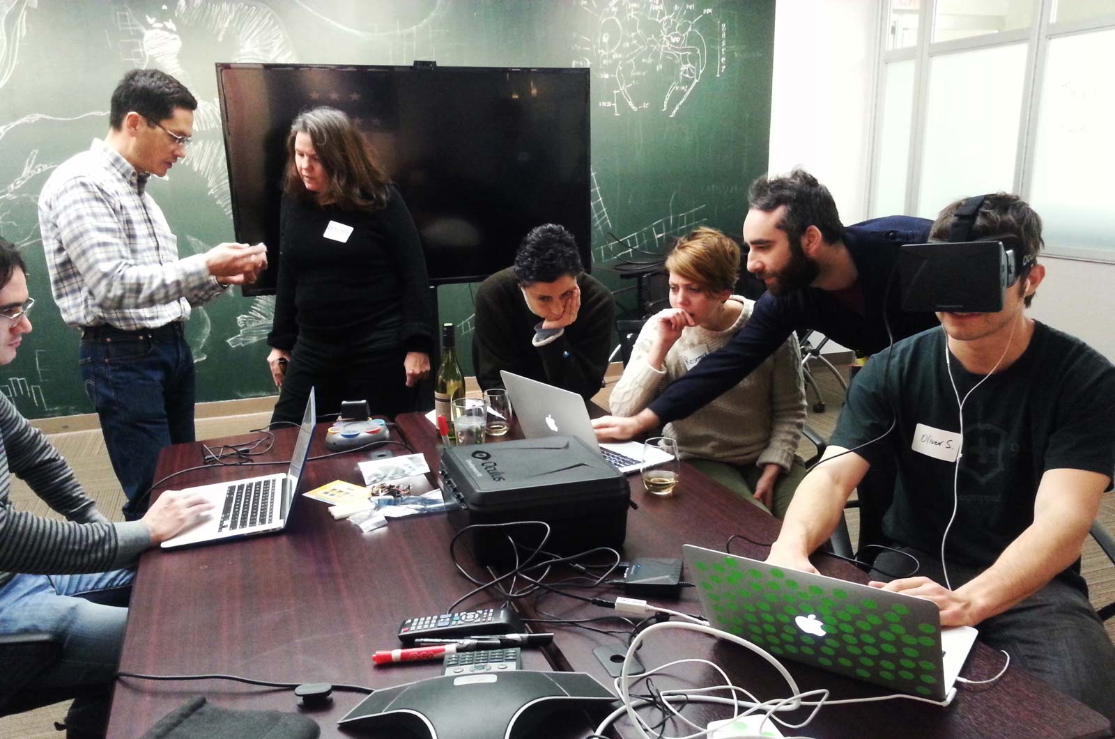 People sitting around a table, looking at computers. One has a VR headset on.