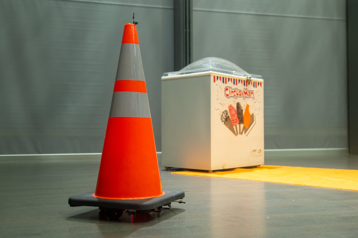 An orange traffic cone sits on a robot with wheels, approaching a “Circus Man” ice cream freezer