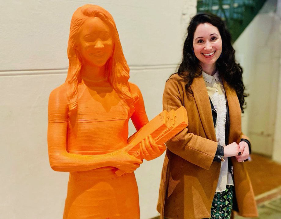 A woman with dark hair smiling next to a 3D printed organge statue of herself