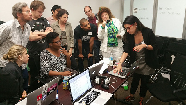 Nine people look on as a workshop participant shows work on a laptop screen