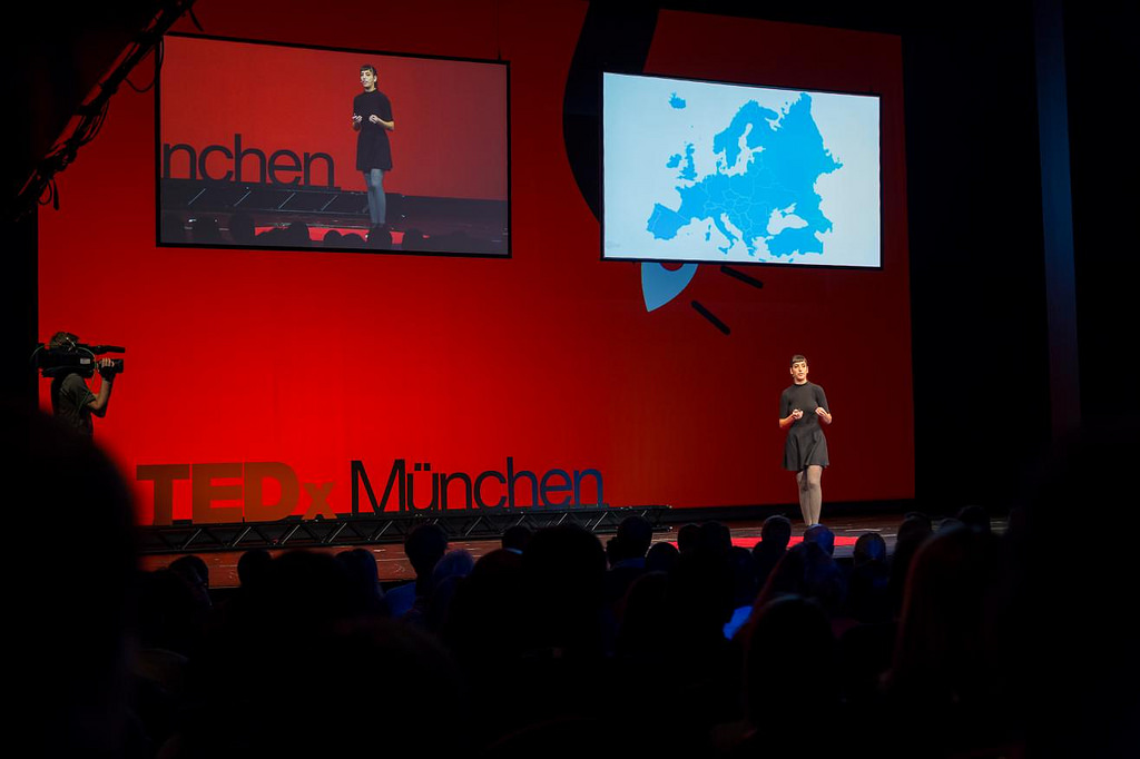 Moon Ribas speaking on a large TEDx stage in red light with a map of Europe displayed on a screen behind her