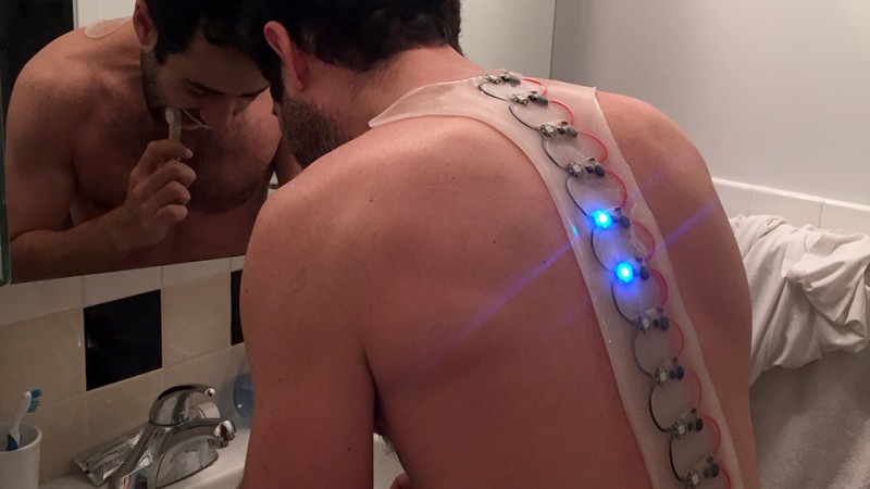 A man seen from behind in a bathroom wearing a synthetic shape down his spine with interlaced electronics