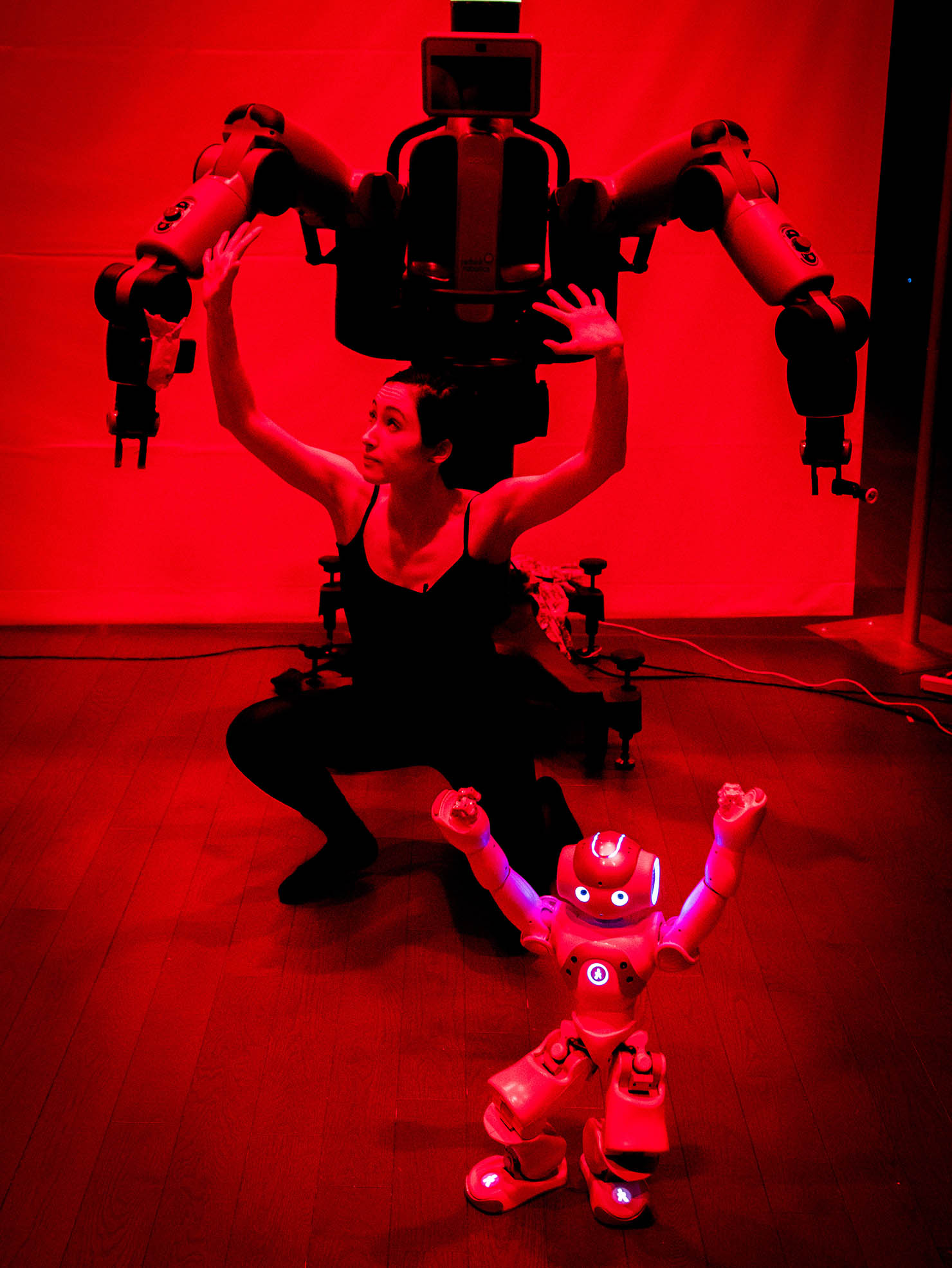 A woman dances with teo humaonoid robots, one small and one large, in a red lit room