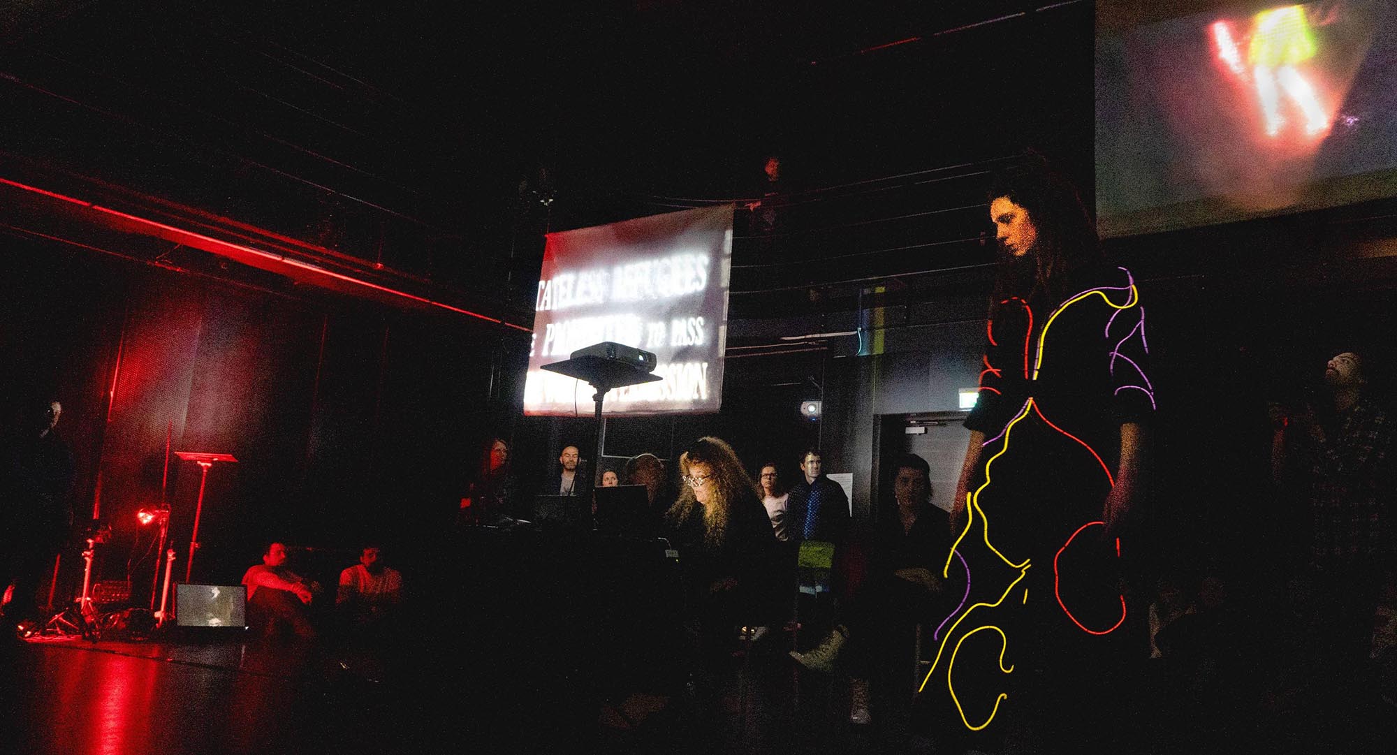 A performer stands in a suit of light in a room with artist Ellen Pearlman and audience members