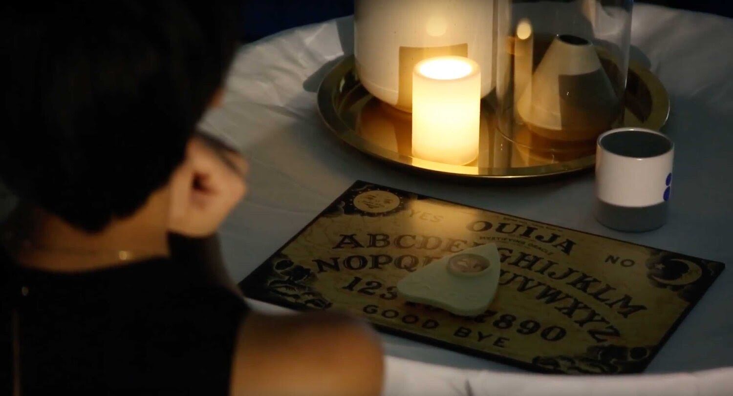 A video still frame looking over a person’s shoulder to see a ouija board