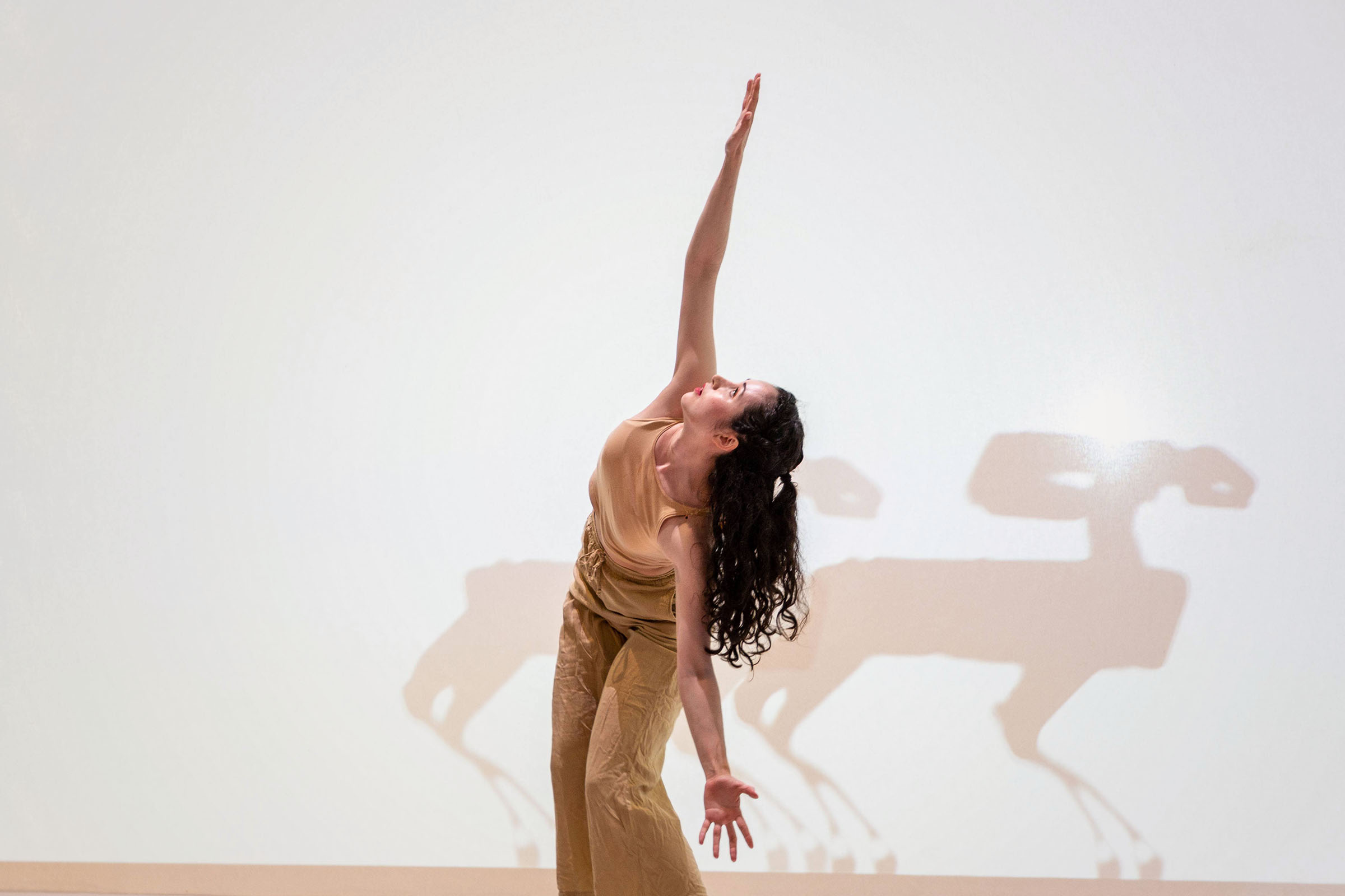 A close-up of a woman dabcing on a stage reaches for the sky with silhouettes of two four-legged animal-like robots behind her