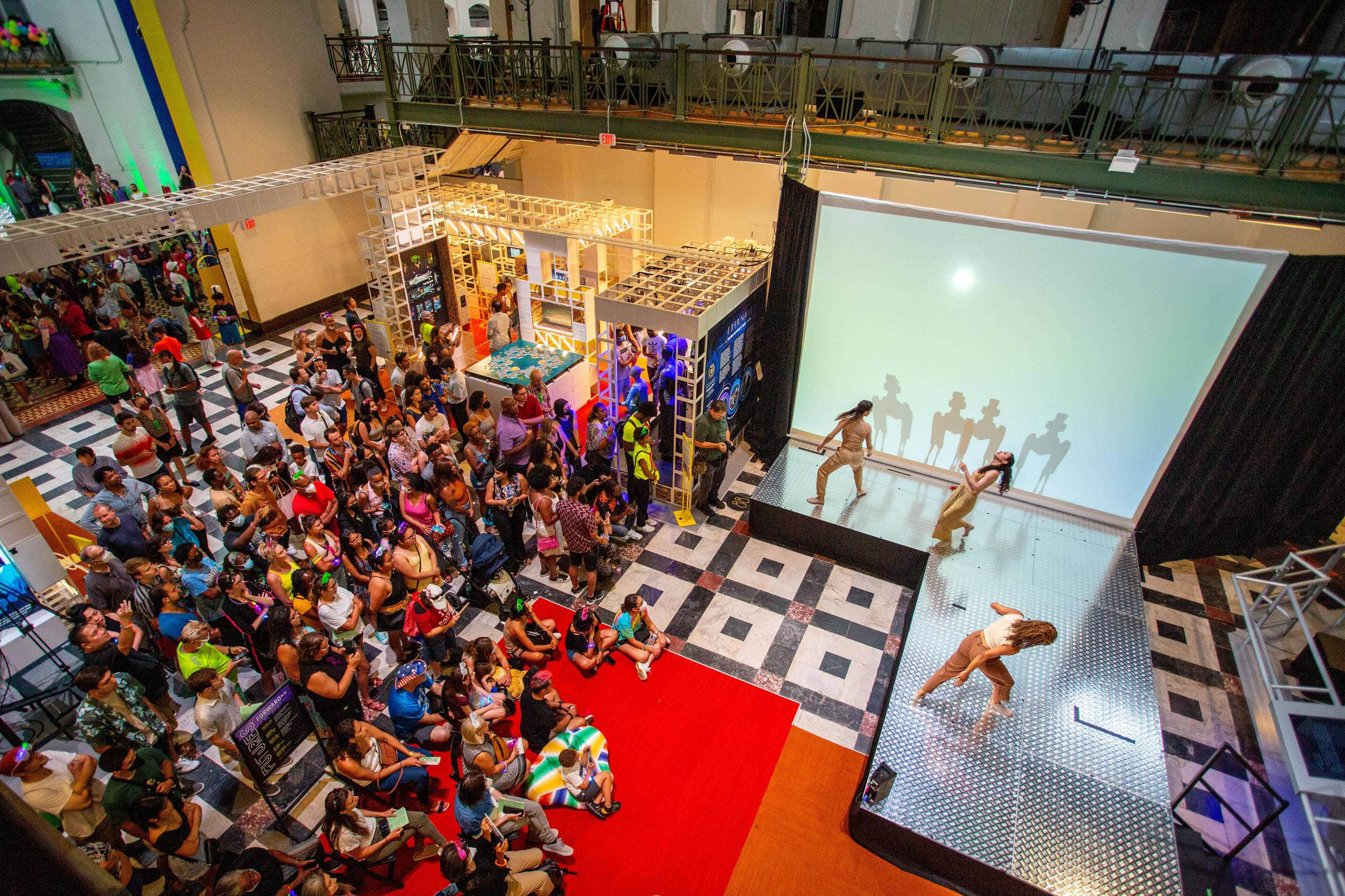 Three dancers moving on a stage in front of a large crowd in an auditorium, with silhouettes of two four-legged animal-like robots behind them