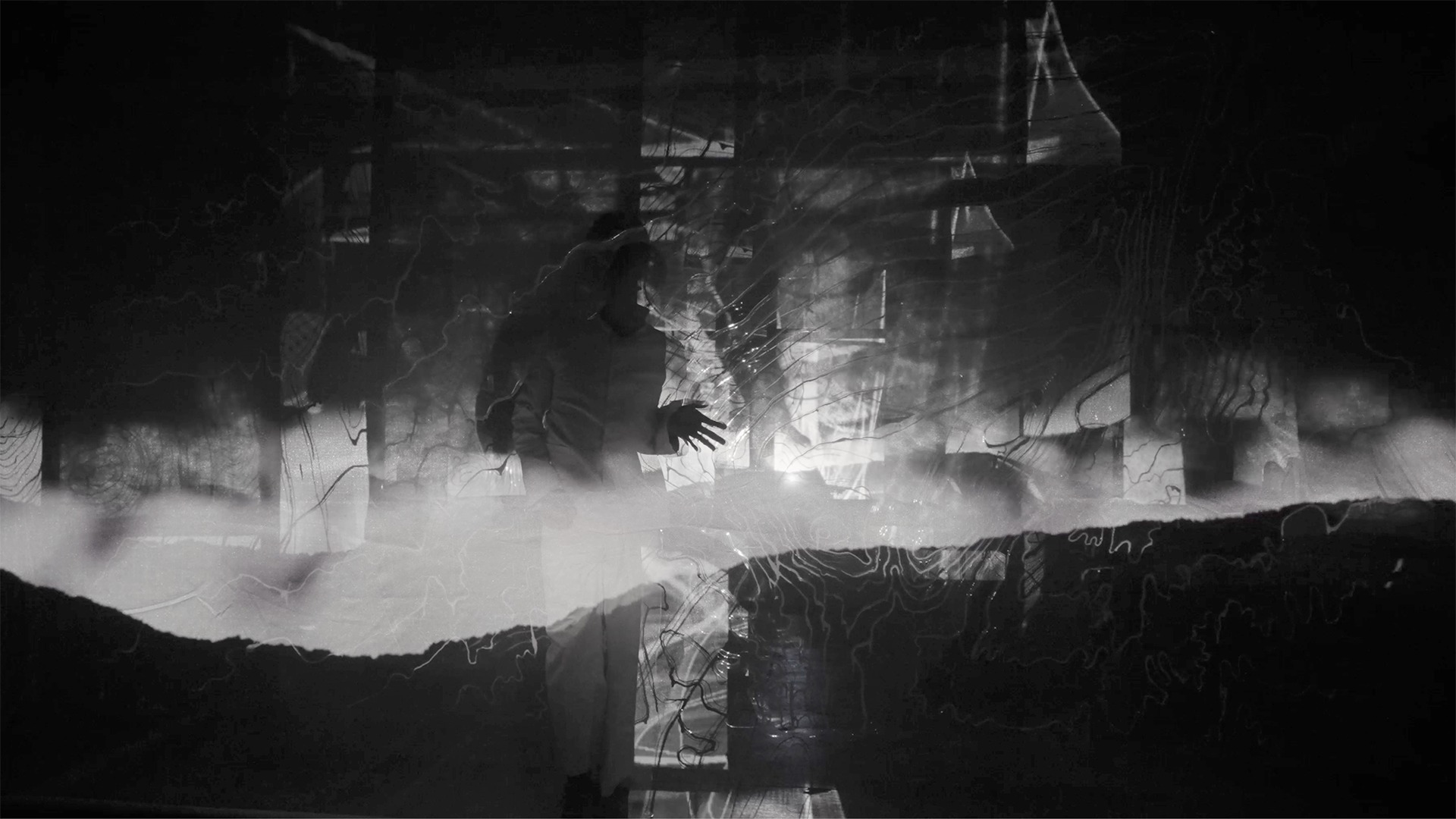 A black and white still of a silhouetted performer reaching through cloudy projected visuals