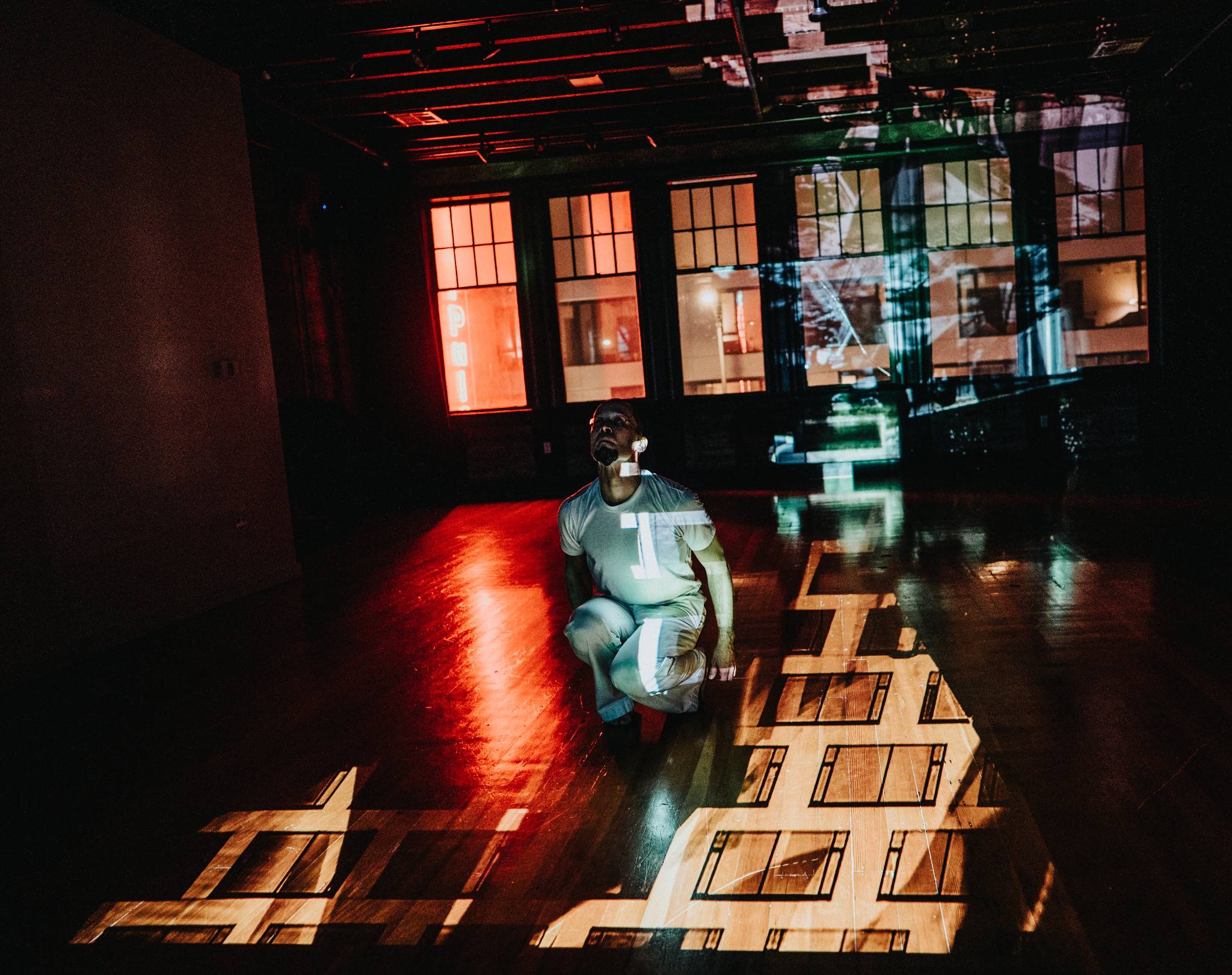 A still image of a male dancer wearing white in a red lit room with abstract white projected visuals