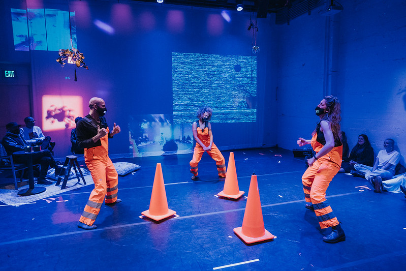 Three performers wearing orange jumpsuits dancing in blue light